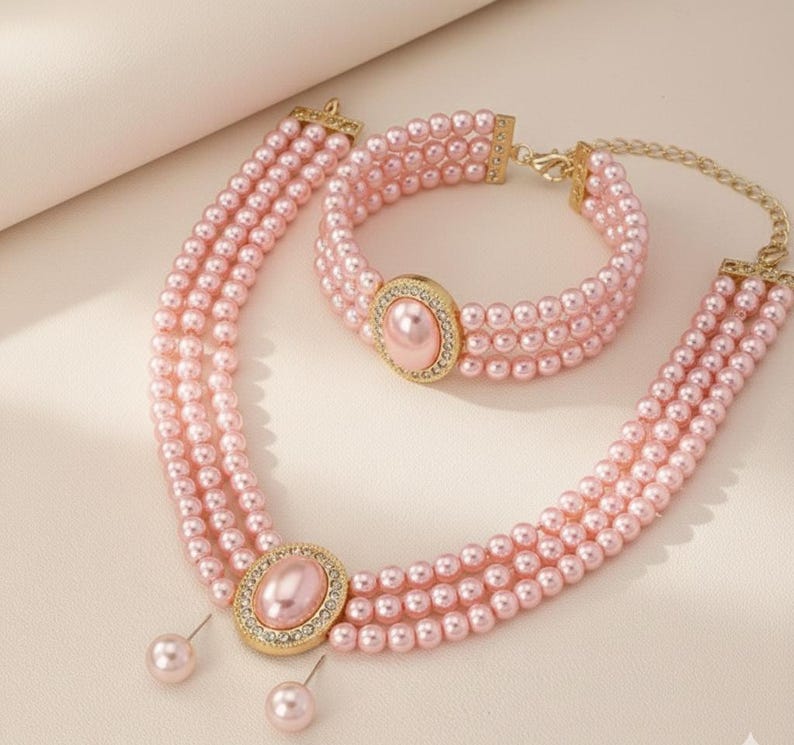 May include: A pink pearl necklace, bracelet, and earring set. The necklace and bracelet feature multiple strands of small, round pearls. Each piece has a gold-colored oval centerpiece with a pink pearl and surrounding clear crystals. The earrings are pink pearl studs.