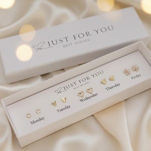 Peut inclure: Une boîte cadeau blanche avec le texte "JUST FOR YOU BEST WISHES" contient six paires de boucles d'oreilles à clous dorées. Chaque paire est conçue pour un jour de la semaine différent, avec diverses formes, dont des cœurs, des papillons et des fleurs.