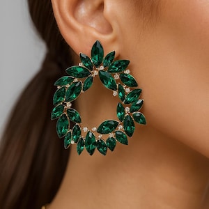 May include: Emerald green hoop earrings. The earrings have a wreath design with marquise-cut green crystals and small clear crystals. The earrings have a gold-tone metal setting. The earrings are designed to be worn on the ear.