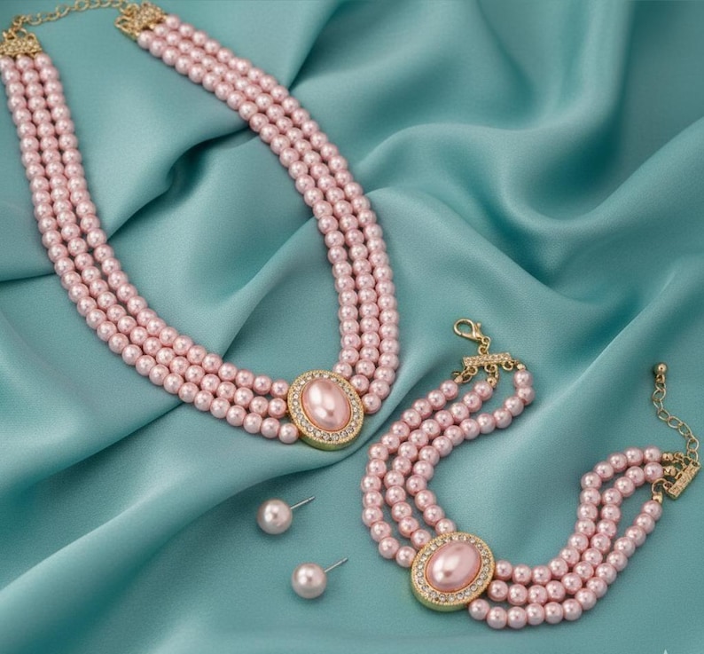 May include: A three-strand pink pearl necklace and bracelet set with gold-tone clasps and oval centerpieces. The centerpieces are adorned with a pink stone and surrounded by small, clear crystals. Matching pearl stud earrings complete the set.