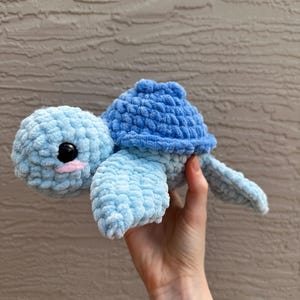 May include: A handmade crochet turtle in shades of light and dark blue. The turtle has a rounded head, a shell, and flipper-like limbs. The turtle's eyes are black, and it has a small pink mouth. The plush toy is held in a hand.