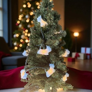 May include: A small artificial Christmas tree decorated with white dog-shaped lights. The tree is placed in front of a blurred background with another Christmas tree and a warm light. The tree is on a red surface.