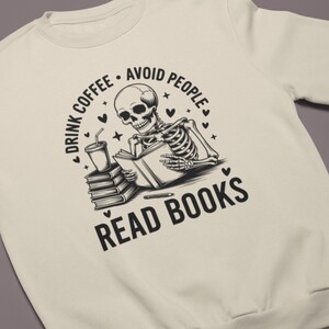 May include: Cream-colored sweatshirt with a black graphic of a skeleton reading a book, holding a coffee cup, and the text "Drink Coffee - Avoid People - Read Books". The design includes hearts and stars.