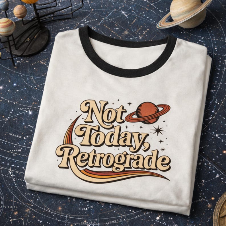 A white t-shirt with a retro-style graphic design and text printed on it, placed on a dark background with a planet model and a starry sky pattern.