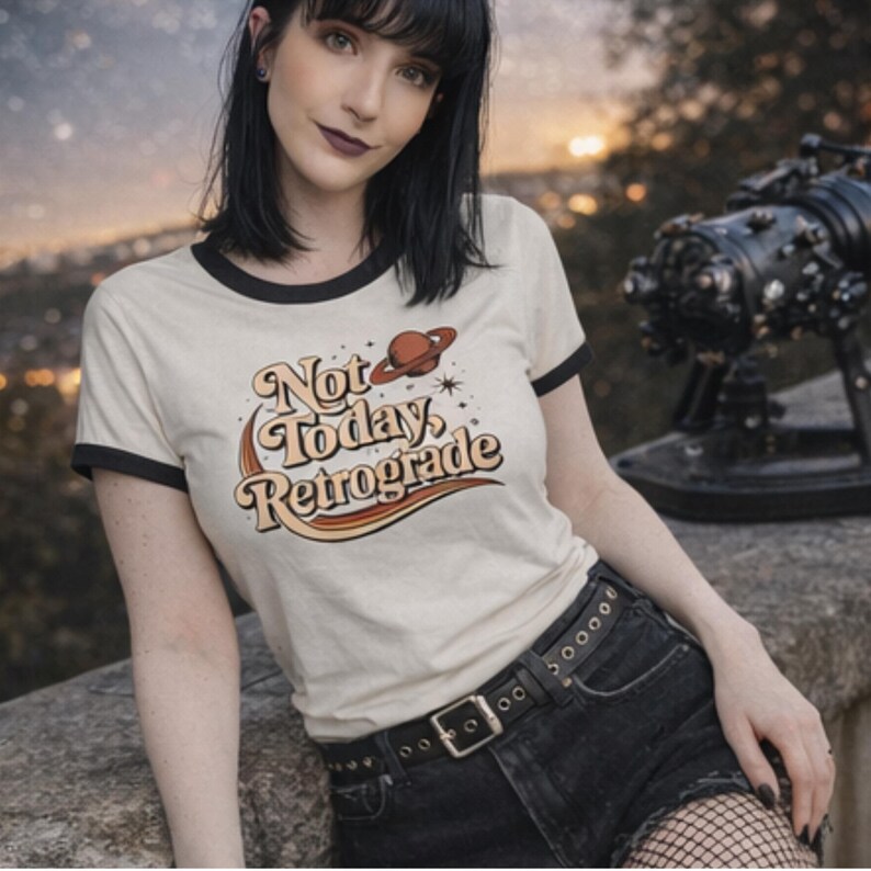 A young woman wearing a white t-shirt with a retro-style graphic and black shorts, posing outdoors against a backdrop of a sunset.