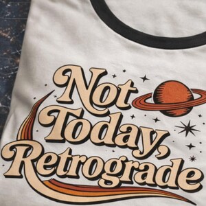 A white t-shirt with a retro-style graphic design featuring the text "Not Today, Retrograde" along with a planet and stars.