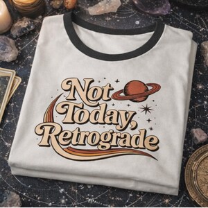 A white t-shirt with a retro-style graphic design that says "Not Today, Retrograde" and features a planet and stars.
