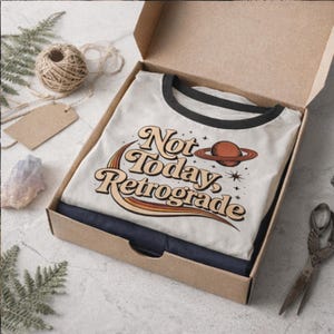 a white t-shirt with a retro-style graphic design, placed in an open cardboard box on a table. There are some twine, scissors, and crystals visible in the scene as well.