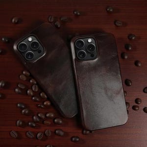 Könnte beinhalten: Zwei dunkelbraune Leder-Handyhüllen mit Kameraausschnitten liegen auf einer Holzoberfläche, auf der Kaffeebohnen verstreut sind. Die Hüllen haben eine strukturierte Oberfläche und sind für ein Smartphone konzipiert.