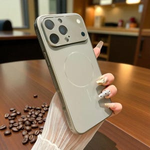 Op de afbeelding: Een lichtgrijze telefoonhoes met een zilveren frame. De hoes heeft een cirkelvormig ontwerp op de achterkant en een cameramodule met meerdere lenzen. De telefoonhoes wordt vastgehouden in een hand met lange, versierde nagels. Koffiebonen liggen verspreid op de tafel.