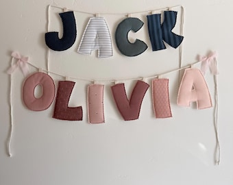 May include: Two fabric letter banners hanging on a wall. The top banner spells JACK in navy, gray striped, solid gray, and navy striped fabric. The bottom banner spells OLIVIA in various shades of pink fabric with small light pink bows.