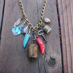 May include: A gold chain necklace with a variety of charms, including a turquoise tooth, a red coral piece, a silver shell, a silver bell, a silver key, and a small skull charm.