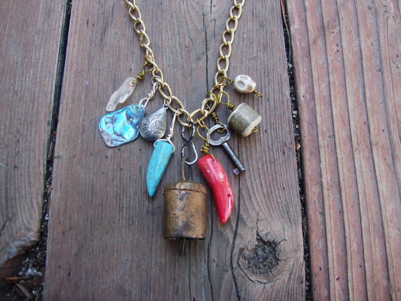 May include: A gold chain necklace with a variety of charms, including a turquoise tooth, a red coral piece, a silver shell, a silver bell, a silver key, and a small skull charm.