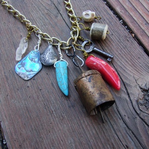 May include: A gold chain necklace with multiple charms, including a turquoise pendant, a red coral pendant, a silver pendant with a floral design, a small silver bell, a key, a bone charm, and a white crystal charm.