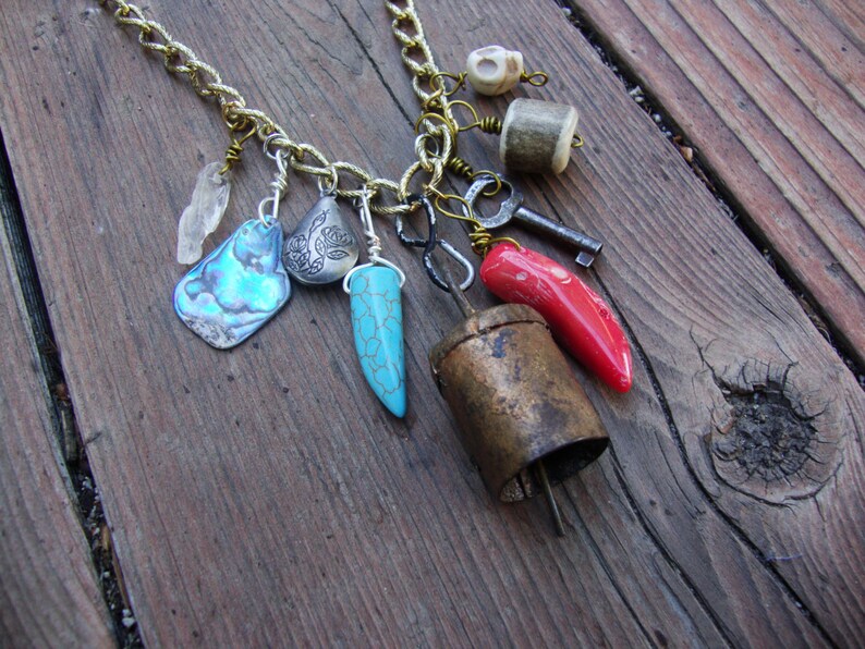 May include: A gold chain necklace with multiple charms, including a turquoise pendant, a red coral pendant, a silver pendant with a floral design, a small silver bell, a key, a bone charm, and a white crystal charm.