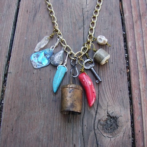 May include: A gold chain necklace with a variety of charms, including a turquoise tooth, a red coral tooth, a small bell, a key, a shell, a crystal, and a bone.