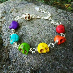 May include: A colorful bracelet featuring skull beads in various colors, including purple, turquoise, green, yellow, orange, and red. The skulls are strung together with silver-colored wire and chain. The bracelet is displayed on a textured, gray surface.