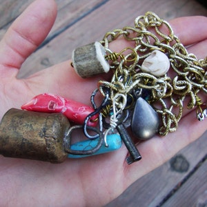May include: A collection of charms and pendants, including a brass bell, a turquoise stone, a red coral, a bone charm, a silver key, a silver chain, and a black stone pendant.