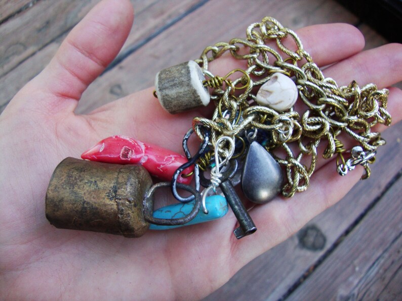 May include: A collection of charms and pendants, including a brass bell, a turquoise stone, a red coral, a bone charm, a silver key, a silver chain, and a black stone pendant.