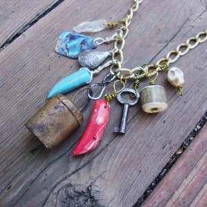 May include: A gold chain necklace with multiple charms, including a bell, a turquoise stone, a red coral, a key, a bone, and a shell.