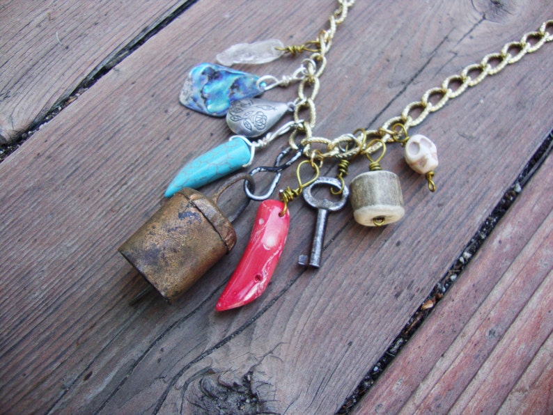 May include: A gold chain necklace with multiple charms, including a bell, a turquoise stone, a red coral, a key, a bone, and a shell.