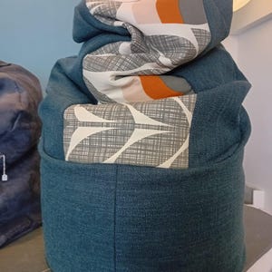 May include: A teal beanbag chair with a patterned top. The top has a design with white, orange, and grey shapes. The beanbag has a cylindrical shape and is made of a textured fabric.