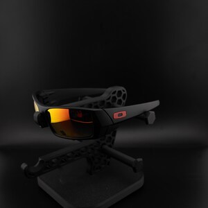 Pode incluir: &Oacute;culos de sol pretos com lentes espelhadas laranja s&atilde;o exibidos em um suporte preto. Os &oacute;culos t&ecirc;m uma arma&ccedil;&atilde;o retangular com um logotipo vermelho na haste. As lentes refletem um tom quente alaranjado.