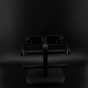 May include: Black sunglasses on a black display stand. The sunglasses have a rectangular frame and dark lenses. The stand features a square base and a vertical support with horizontal bars for the glasses.