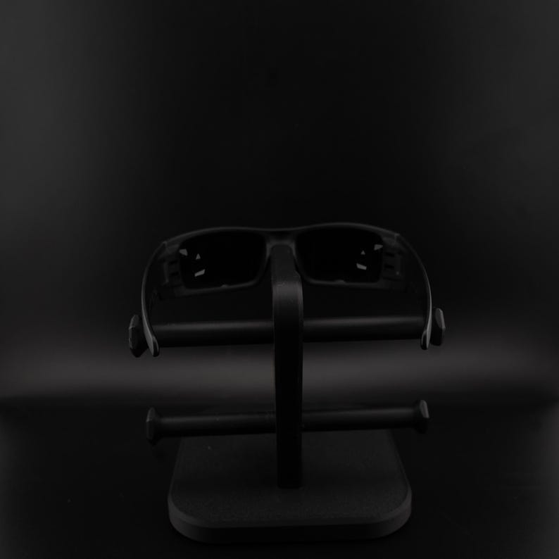 May include: Black sunglasses on a black display stand. The sunglasses have a rectangular frame and dark lenses. The stand features a square base and a vertical support with horizontal bars for the glasses.