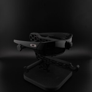 May include: Black sunglasses with dark lenses, presented on a black stand. The sunglasses feature a rectangular frame with a small logo. The stand has a geometric design, set against a black backdrop.