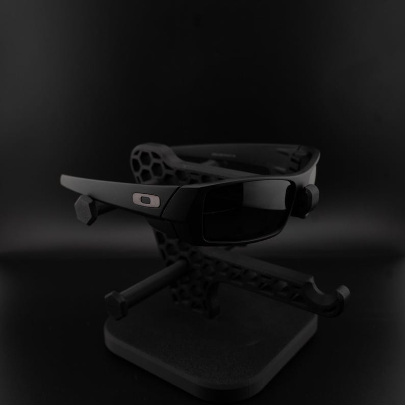 May include: Black sunglasses with dark lenses, presented on a black stand. The sunglasses feature a rectangular frame with a small logo. The stand has a geometric design, set against a black backdrop.