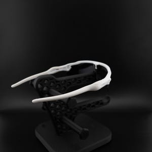 May include: White sports sunglasses with a sleek, modern design. The sunglasses have a white frame and dark lenses. They are displayed on a black stand against a black background, highlighting their sporty aesthetic.