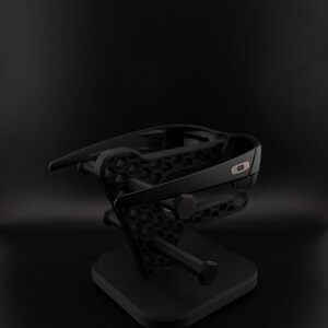 May include: Black sunglasses displayed on a black stand. The sunglasses have a dark frame and lenses. The stand has a geometric pattern and a square base, creating a minimalist aesthetic. The stand is likely made of plastic.