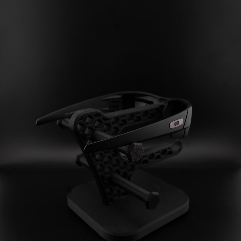 May include: Black sunglasses displayed on a black stand. The sunglasses have a dark frame and lenses. The stand has a geometric pattern and a square base, creating a minimalist aesthetic. The stand is likely made of plastic.