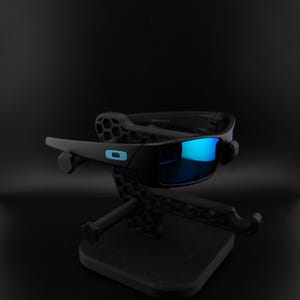 May include: Black sunglasses with blue mirrored lenses are displayed on a black stand. The sunglasses have a rectangular frame with a small blue logo on the side. The stand has a geometric design.