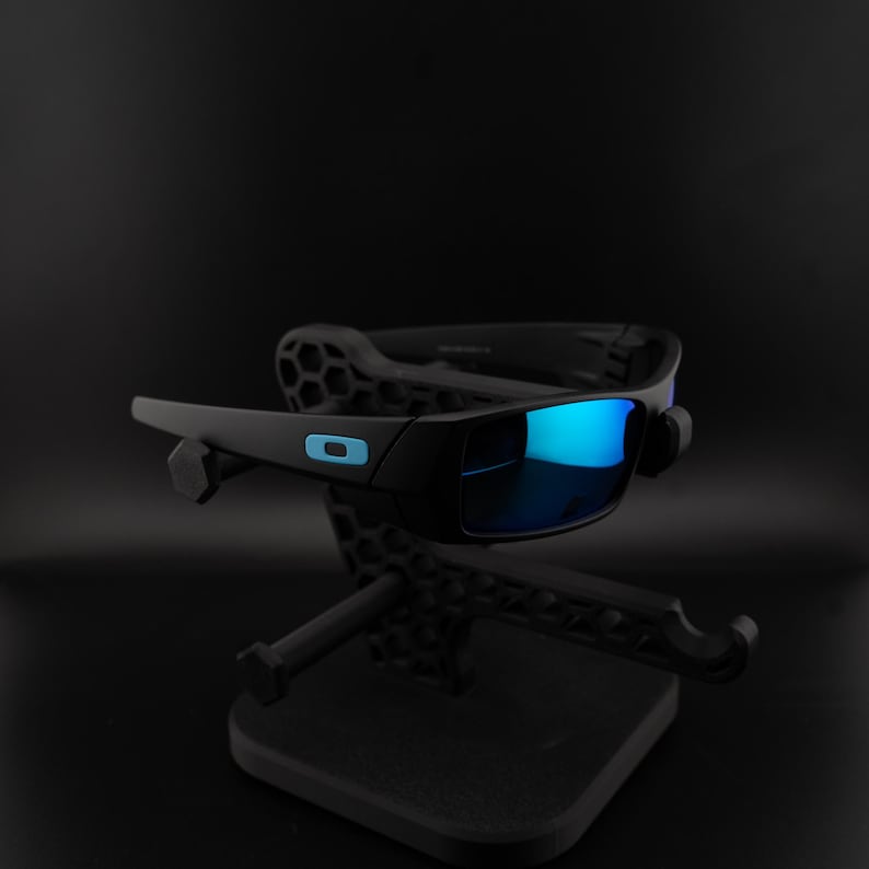 May include: Black sunglasses with blue mirrored lenses are displayed on a black stand. The sunglasses have a rectangular frame with a small blue logo on the side. The stand has a geometric design.
