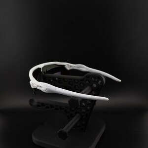May include: White sports sunglasses with a black lens and a white frame. The sunglasses are displayed on a black stand against a black background. The Oakley logo is visible on the side of the frame.