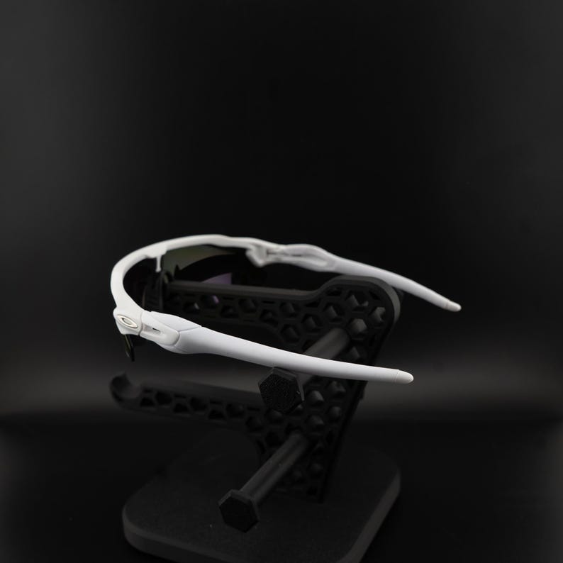 May include: White sports sunglasses with a black lens and a white frame. The sunglasses are displayed on a black stand against a black background. The Oakley logo is visible on the side of the frame.