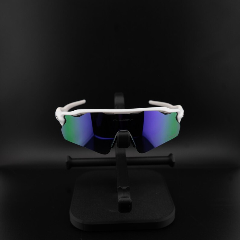 May include: White sports sunglasses with mirrored blue and green lenses. The sunglasses are displayed on a black stand against a black background. The frames are white and have a sleek, modern design.