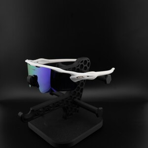 May include: White-framed sunglasses with blue and green lenses are displayed on a black stand. The sunglasses have a modern, sporty design. The stand is also black and has a geometric pattern.