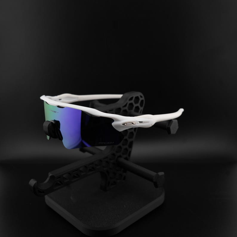May include: White-framed sunglasses with blue and green lenses are displayed on a black stand. The sunglasses have a modern, sporty design. The stand is also black and has a geometric pattern.