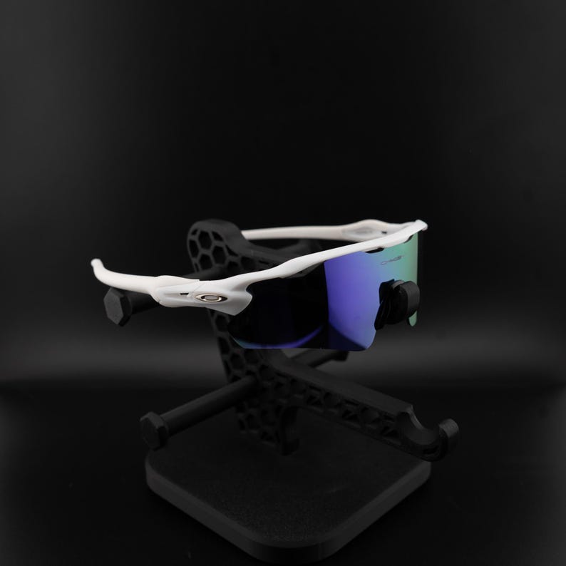 May include: White-framed sunglasses with blue and green lenses rest on a black display stand. The sunglasses have a sporty design, and the stand has a geometric pattern. The background is a solid black.
