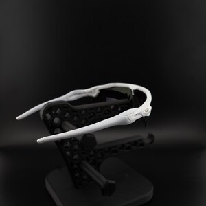 May include: White sports sunglasses with a sleek design, resting on a black display stand. The sunglasses have a curved frame and dark lenses, suitable for outdoor activities. The stand has a geometric pattern.