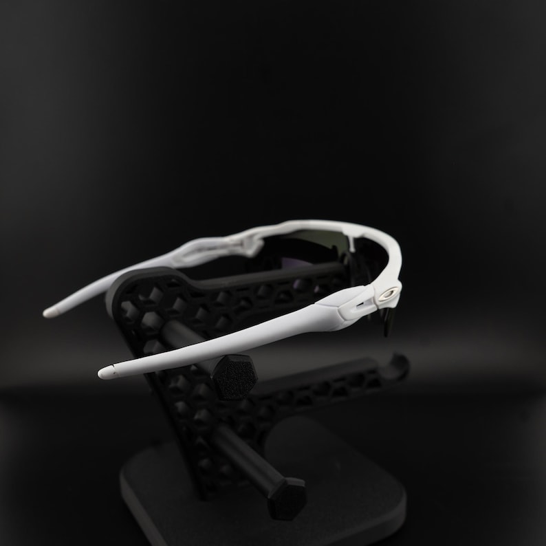 May include: White sports sunglasses with a sleek design, resting on a black display stand. The sunglasses have a curved frame and dark lenses, suitable for outdoor activities. The stand has a geometric pattern.