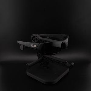 May include: Black sunglasses with dark lenses are displayed on a black stand. The sunglasses feature a rectangular frame with a silver detail. The stand has a geometric pattern and a square base, set against a dark backdrop.