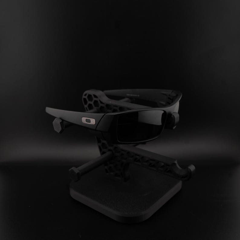 May include: Black sunglasses with dark lenses are displayed on a black stand. The sunglasses feature a rectangular frame with a silver detail. The stand has a geometric pattern and a square base, set against a dark backdrop.