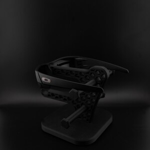 May include: Black sunglasses displayed on a black stand. The sunglasses have a modern design with a small, silver logo. The stand has a geometric pattern and a square base, suitable for displaying eyewear.