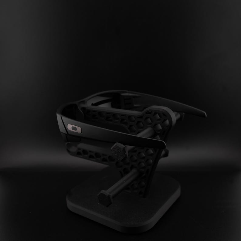 May include: Black sunglasses displayed on a black stand. The sunglasses have a modern design with a small, silver logo. The stand has a geometric pattern and a square base, suitable for displaying eyewear.