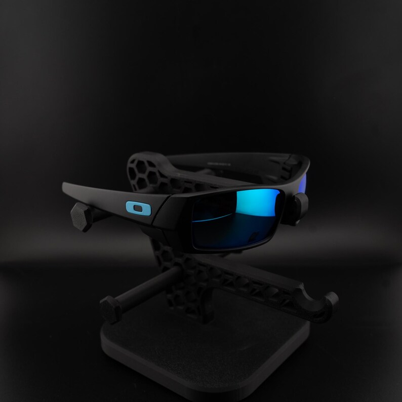 May include: Black sunglasses with blue reflective lenses are displayed on a black stand. The sunglasses have a rectangular frame with a small, light blue logo on the side. The lenses reflect a bright blue color.