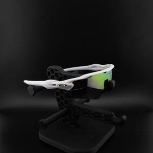 May include: White sports sunglasses with iridescent lenses are displayed on a black stand. The sunglasses have a sleek, modern design with a white frame and a prominent logo on the side. The lenses reflect a mix of green and purple hues.